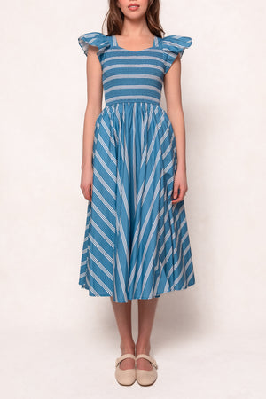 A woman wears the Ivy City Co Adelaide Dress in Blue, a cotton midi dress with blue and white stripes, flutter sleeves, a smocked bodice, and pockets. She stands facing forward against a plain light background in beige Mary Jane shoes.