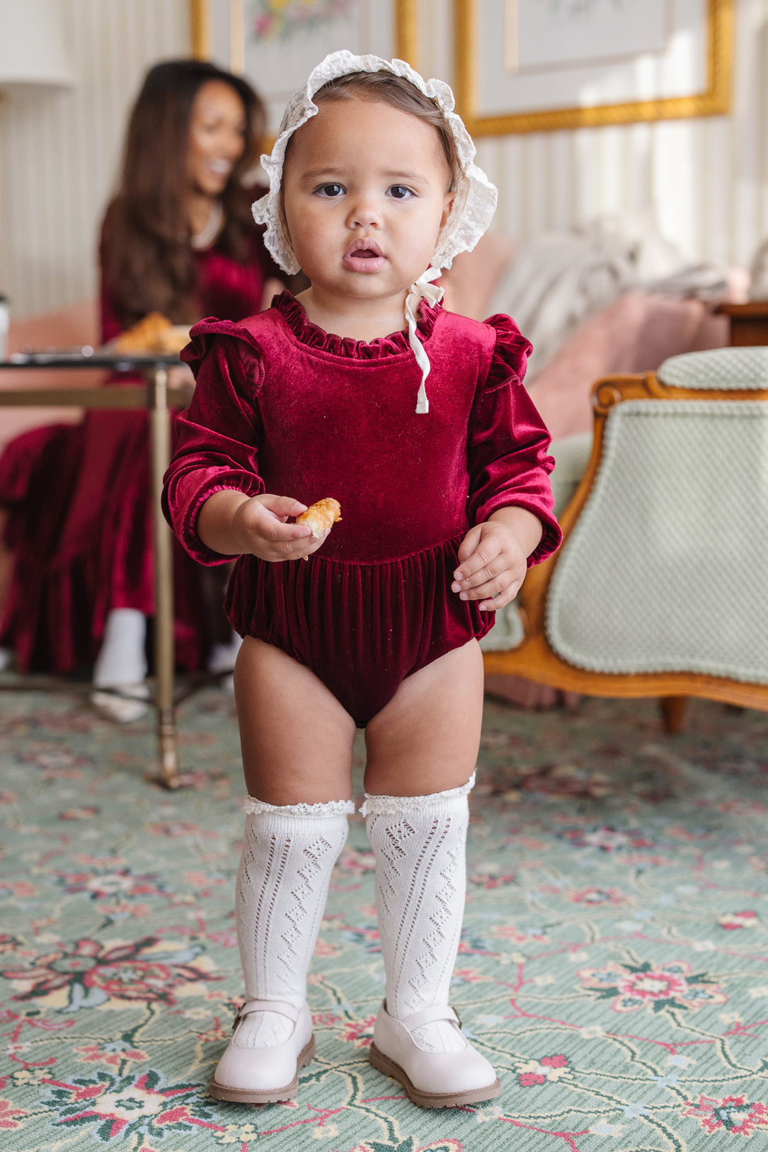 baby in a red velvet romper and white socks in a living room