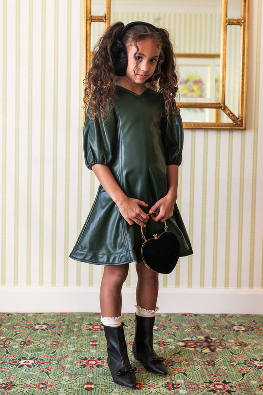 A girl wearing a green vegan leather mini dress with a v neckline and long balloon sleeves. She is also wearing white tights and black boots, carrying a small black purse.