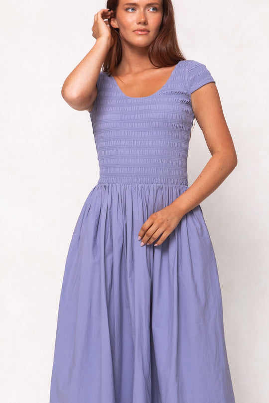 A woman wearing the Ivy City Co Marlow Cap Sleeve Dress in Blue, featuring a fitted bodice and flowing skirt, stands against a plain light background, touching her hair with one hand.