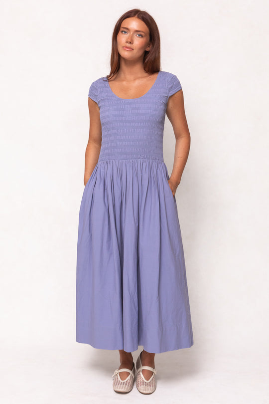 A woman with long brown hair wears the Ivy City Co Marlow Cap Sleeve Dress in Blue, an ankle-length, nursing-friendly dress with a smocked bodice and pockets, paired with white woven flats, against a plain light background.