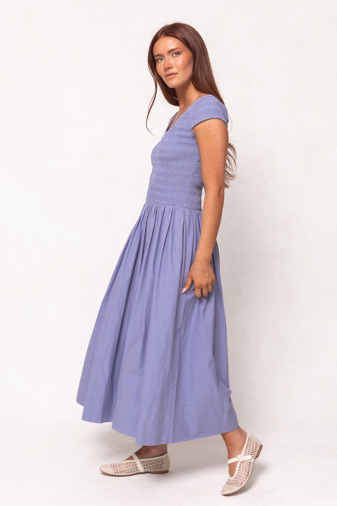 A woman with long brown hair stands sideways against a plain light background, wearing the Ivy City Co Marlow Cap Sleeve Dress in Blue and white woven flats.
