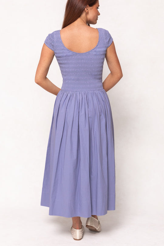 A woman with long brown hair wears the Ivy City Co Marlow Cap Sleeve Dress in Blue, featuring short sleeves, a smocked bodice, an ankle-length skirt, and a nursing-friendly scoop back. She stands turned away in light-colored shoes.