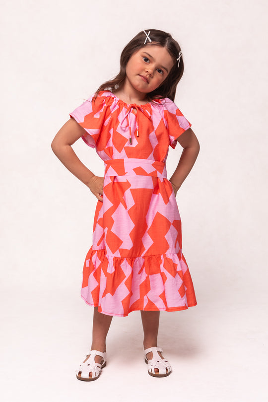 A young girl with long brown hair and hair clips poses with her hands on her hips, wearing the Ivy City Co Mini Haven Dress in Geometric and white sandals against a plain, light background.