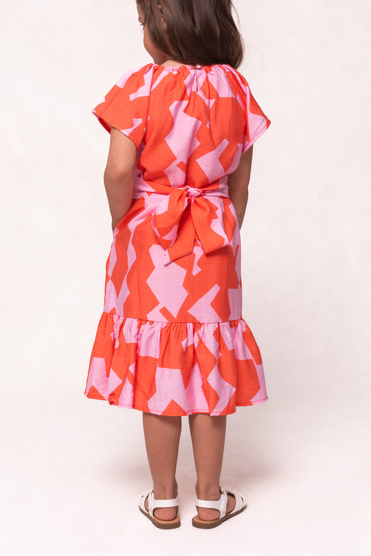 A young child faces away, wearing Ivy City Co’s Mini Haven Dress in Geometric—a pink and orange cotton dress with a ruffled hem and back bow—paired with white sandals against a plain, light background.