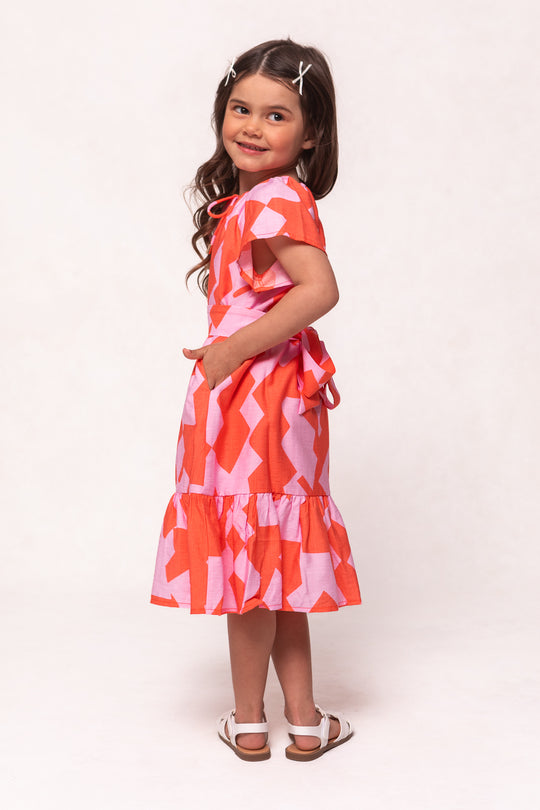 A young girl with long dark hair and white hair clips smiles while wearing the Ivy City Co Mini Haven Dress in Geometric and white sandals, standing sideways with one hand in her pocket against a light background.