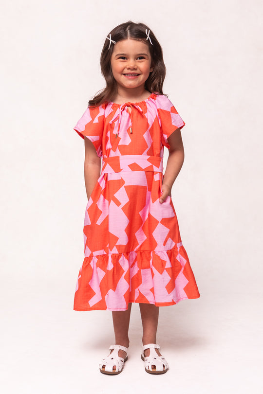 A smiling girl with long brown hair in white clips wears the Ivy City Co Mini Haven Dress in Geometric—a pink and orange cotton dress with ruffled hem and short sleeves—paired with white sandals against a plain light background.