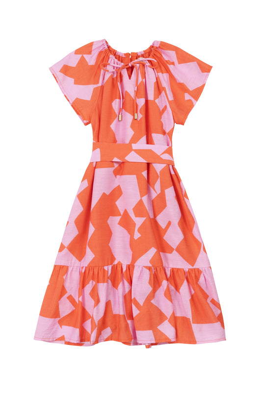 The Ivy City Co Mini Haven Dress in Geometric is a short-sleeve, knee-length cotton dress featuring a pink and orange geometric print, round neckline with drawstring tie, fitted waist, and flared ruffled skirt.