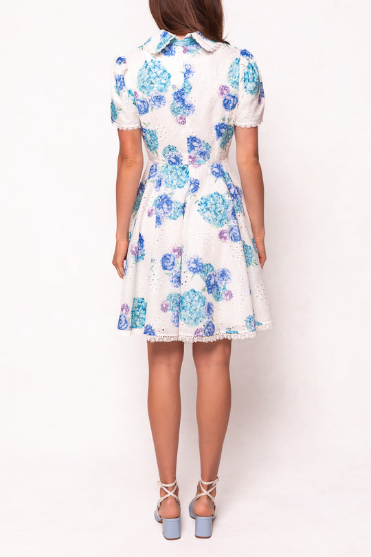 A woman stands facing away in the Ivy City Co Penny Dress in Blue Floral, featuring cotton eyelet fabric, blue and purple floral patterns, a lace hem, and styled with light blue high-heeled sandals against a plain white background.