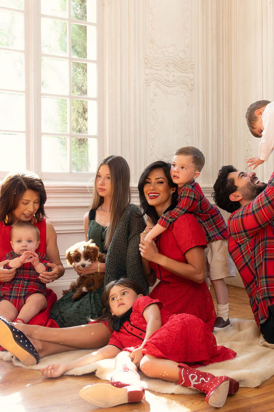 A family dressed in coordinated red and plaid outfits, including the adorable Ivy City Co Baby Madeline Romper in Holiday Plaid as the perfect holiday look for baby, sits together by a sunlit window. Children, adults, and a dog are all gathered closely on the floor, sharing joyful smiles.
