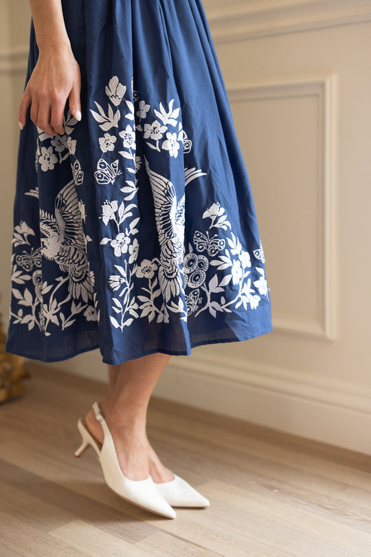 A person stands indoors on wooden flooring wearing the Finch Dress in Blue by Ivy City Co, a deep blue cotton dress decorated with delicate white bird and flower embroidery, complemented by white slingback heels.