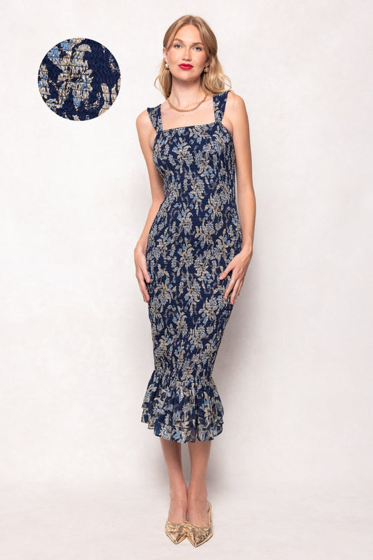 A woman wears the Mira Dress in Gilded Vine by Ivy City Co—a fitted, navy blue midi dress featuring a luxurious floral brocade, smocked bodice, ruffled hem, and shoulder straps—standing against a light background. An inset highlights a close-up of the elegant Gilded Vine patterned fabric.