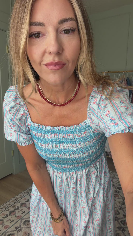 A woman with long hair is standing indoors, looking at the camera while wearing the Ivy City Co Sunday Best Dress in Rosette Linear Stripe—a blue and white floral cotton midi dress with puff sleeves—paired with a red necklace.