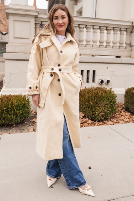 A woman stands outside near bushes and a light-colored building, smiling as she poses on the sidewalk in Ivy City Co’s York Coat in Cream. The long, tailored coat with structured shoulders is paired with blue jeans and pointed cream flats.