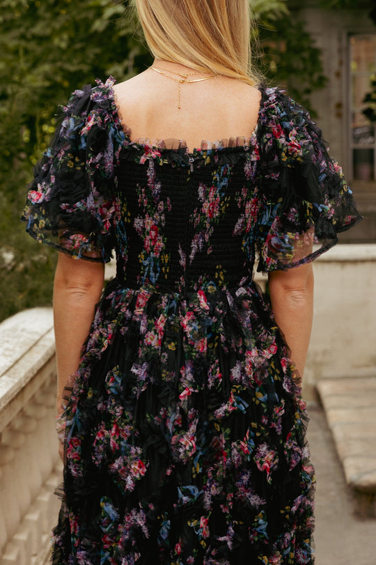 A woman with blonde hair stands outdoors wearing the Florentina Dress in Obsidian Garden by Ivy City Co, a stunning black tulle gown adorned with colorful floral patterns, ruffled details, and sheer sleeves. Photographed from behind to highlight its exquisite texture and vibrant design, this dress is a beautiful maternity-friendly option for an evening out.