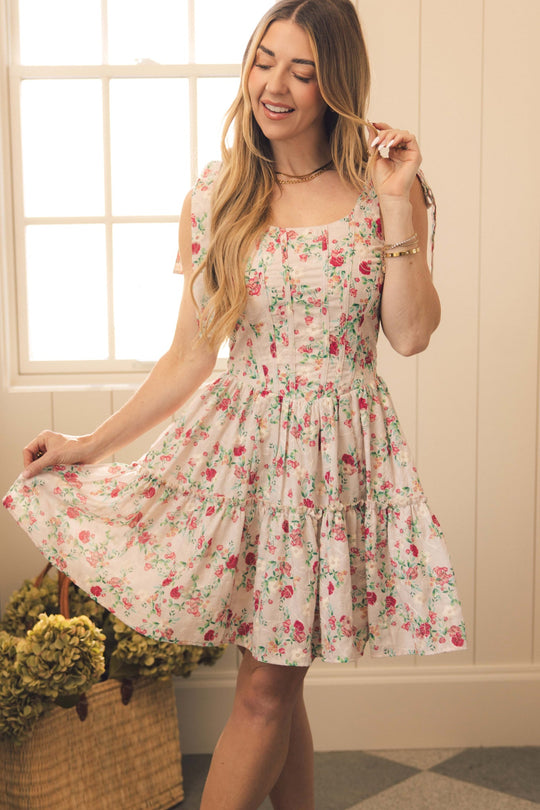 A woman wearing the Helene Dress in Vibrant Chintz by Ivy City Co stands indoors, smiling as she holds out her skirt. Sunlight streams through a window behind her, and a basket filled with green and yellow flowers rests on the floor.