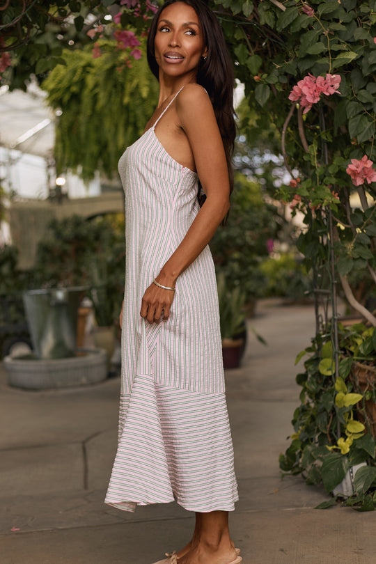 A woman wearing the Marbella Dress in Green & Pink Pinstripe by Ivy City Co stands sideways in a lush garden. Pink flowers and greenery surround her as she smiles and gazes into the distance, highlighting the effortless elegance of her striped midi dress.