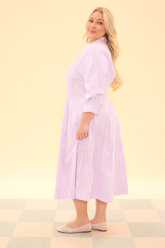 A woman with long blonde hair stands sideways, smiling, and wearing the Ivy City Co Cameron Dress in Lavender—a nursing-friendly, pinstripe midi dress with long sleeves—paired with light-colored flats. The background is a soft pale yellow, and the floor features a light checker pattern.