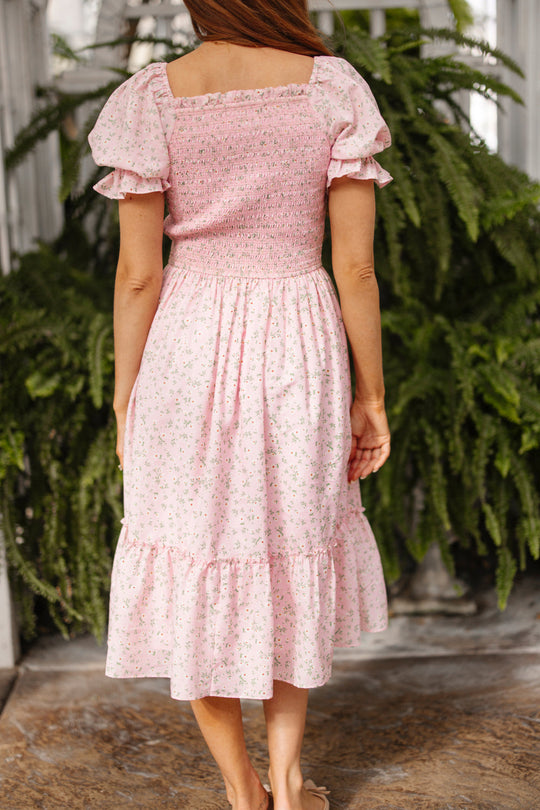 A woman with long hair stands facing away, wearing the Ivy City Co Valencia Dress in Pink Ditsy Floral. This maternity-friendly, short-sleeve dress features a smocked bodice and ruffled hem. Lush green ferns fill the background.