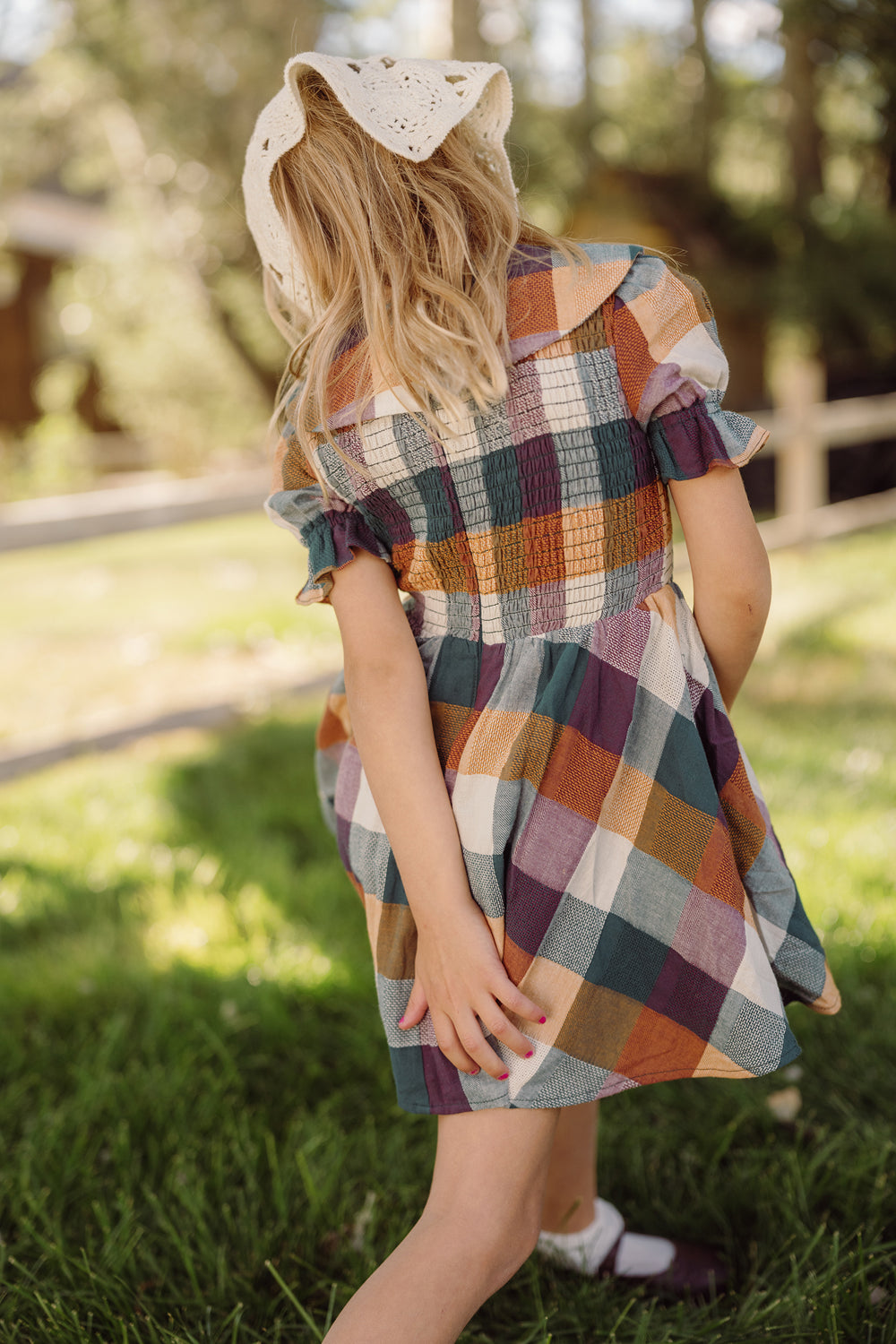 A young girl in the Mini Alice Dress in Maize Gingham by Ivy City Co, featuring a smocked bodice and puff sleeves, stands outside on green grass with her back to the camera, one hand behind her back as sunlight filters through the trees.