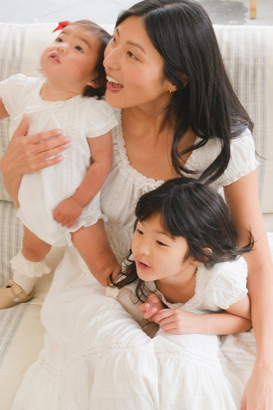 A woman and two young children in white outfits sit closely on a sofa, smiling and looking in different directions. One of the girls wears the Ivy City Co Mini Palermo Dress in White, featuring a smocked bodice and puff sleeves that add to the scene’s timeless charm.