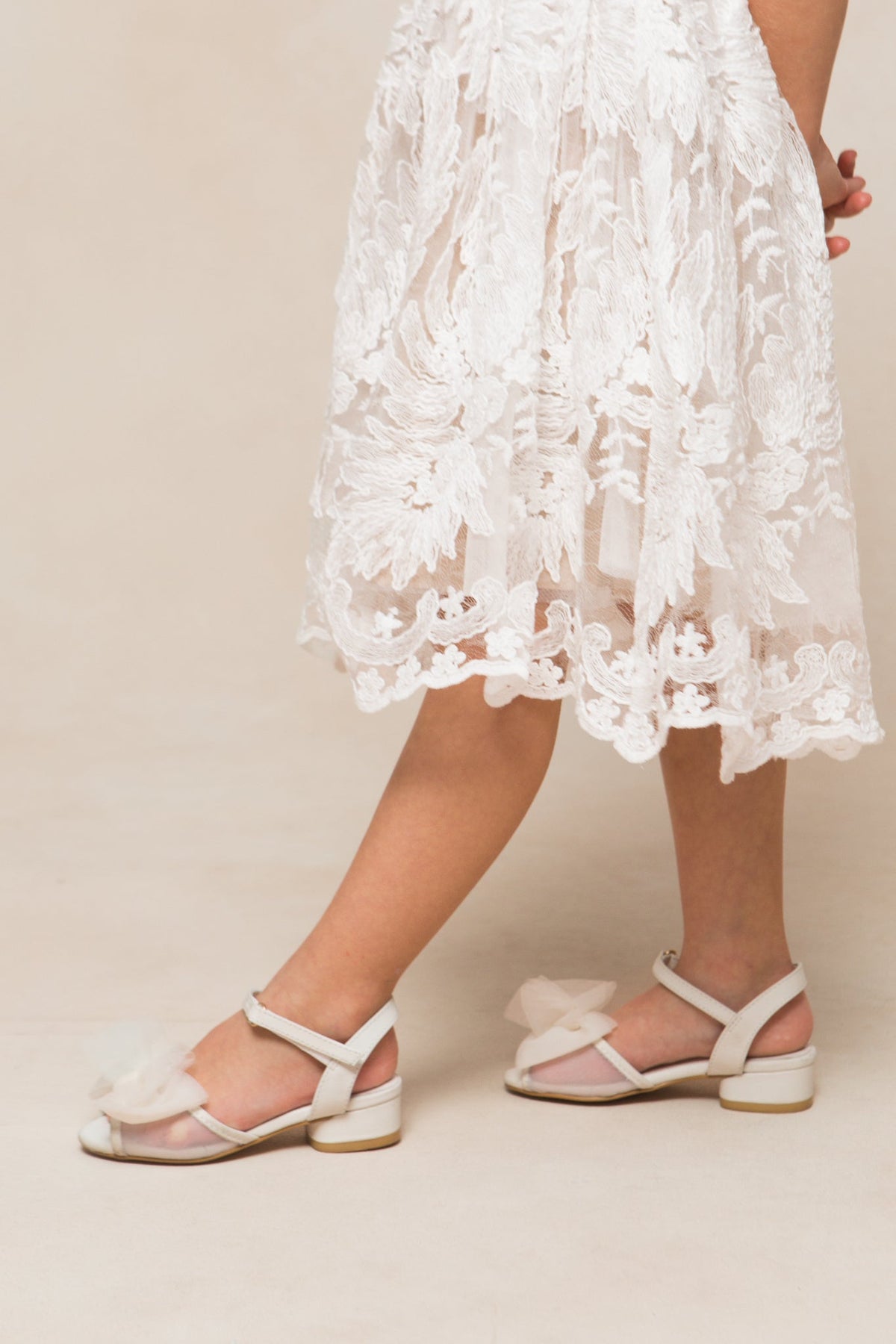 Wearing a white lace dress and comfortable Mini Ivy Bow Heel in White by Ivy City Co—featuring a delicate tulle bow detail—a person stands against a neutral background, with only the lower half of their body visible.