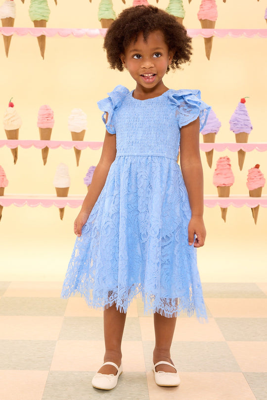 A young girl wearing the Ivy City Co Mini Hattie Dress in Cornflower Blue Lace and white shoes smiles while standing indoors. Pastel ice cream cones are arranged in rows on a light yellow wall behind her, creating a sweet setting perfect for mommy-and-me matching moments.