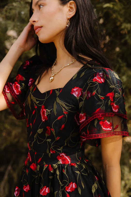 A woman stands outdoors amidst greenery, wearing the Ivy City Co Flora Dress in Romantic Roses, featuring embroidered red roses and a black mesh overlay. She touches her hair and accessorizes with gold jewelry, including a heart pendant necklace.