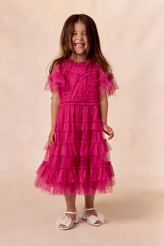 A young girl smiles in the Ivy City Co Mini Whimsical Dress in Fuchsia, featuring flutter sleeves and a layered tulle skirt, paired with white sandals against a light beige background.