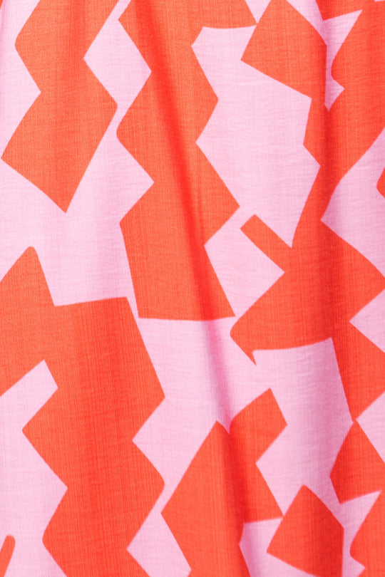 A close-up of the Haven Dress in Geometric by Ivy City Co, showcasing fabric with an abstract zigzag pattern in vibrant orange and soft pink hues. The design features bold, angular shapes that overlap for a striking visual effect.