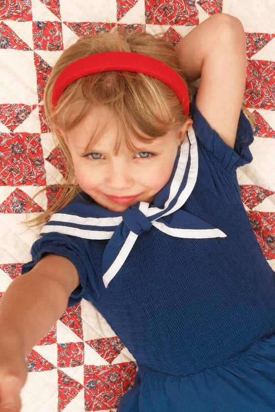 A young girl with blonde hair in an Ivy City Co Mini Alice Dress in Navy and a red headband smiles while lying on a patterned quilt. She stretches one arm toward the camera, showcasing her adorable smocked dress.