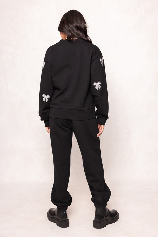 A person stands with their back to the camera, wearing a black sweatshirt paired with Ivy City Co’s Micah Jogger Sweatpants in Black. White bow designs adorn the sleeves, echoed by shimmering rhinestone bow accents that add a subtle sparkle. Their long dark hair cascades over the outfit, complemented by black boots, while a plain light-colored background ensures all attention remains on their stylish Ivy City Co ensemble.