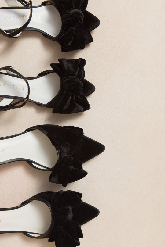 Four pairs of the Ivy City Co Mini Eloise Bow Heel in Black, featuring black velvet pointed toes and large bows, are arranged in a vertical line on a light beige surface.