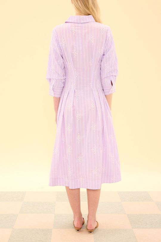 A woman stands with her back to the camera, wearing the Ivy City Co Cameron Dress in Lavender—a midi-length shirt dress with long sleeves and white vertical stripes. The elegant design is nursing friendly and set against a light, neutral backdrop with a checkered floor.