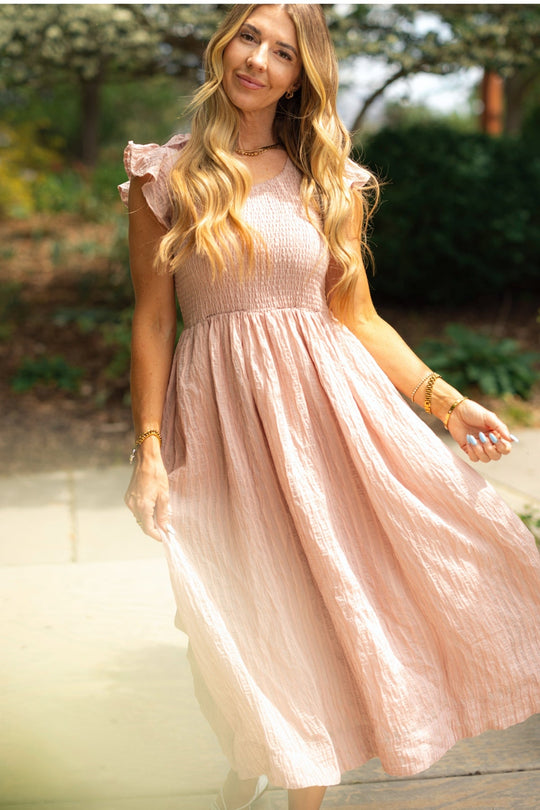 A woman with long, wavy blonde hair stands outdoors on a sunny day amid greenery, smiling gently as she wears the Ivy City Co Hattie Dress in Pink Stripes paired with gold jewelry.