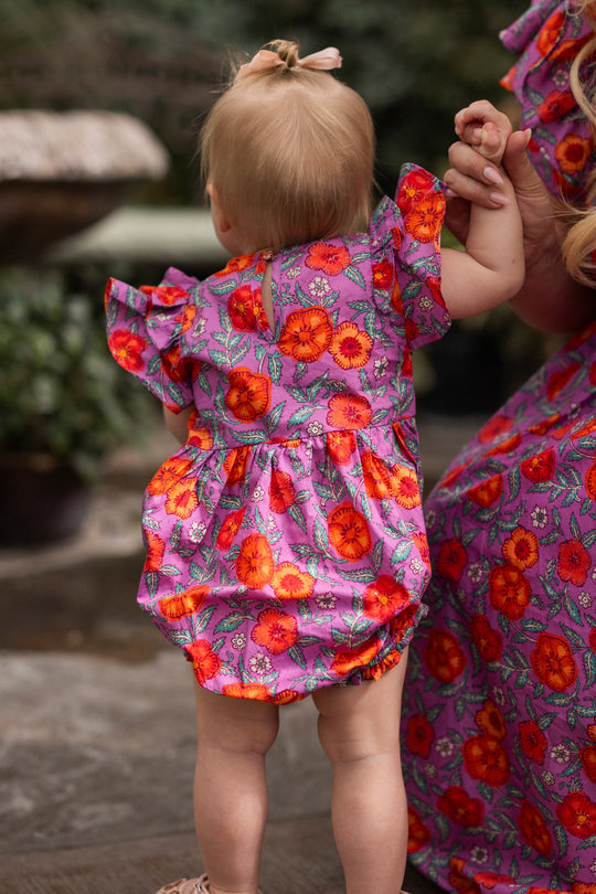 A baby stands near a stone fountain and lush greenery, holding an adult’s hand as they both sport matching bright pink floral patterns. The little one wears the adorable Baby Hattie Romper in Thistle Flower Poppy Woodblock by Ivy City Co—a charming cotton romper ideal for warm days outdoors. With light hair tied in a bow, the baby enjoys stylish moments together with their loved one.