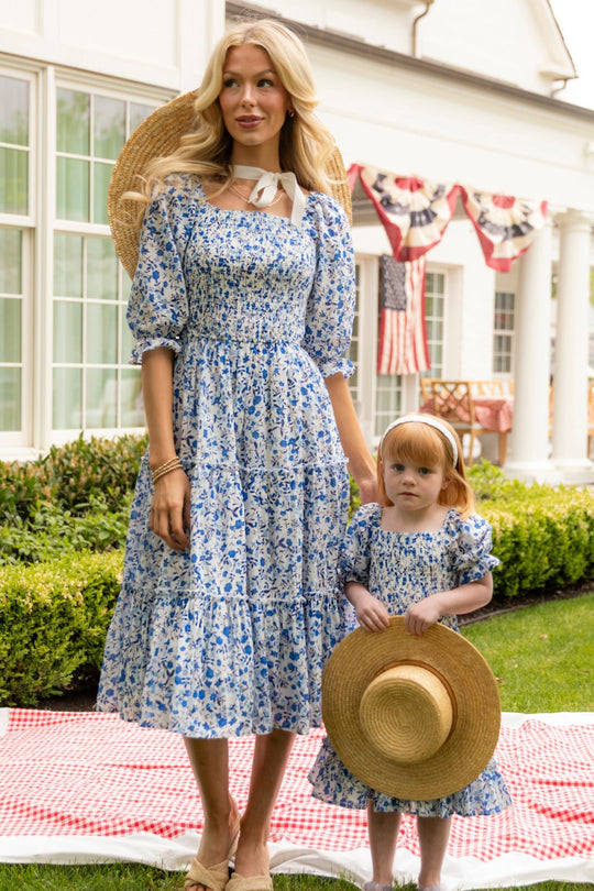 A woman and a young girl, both dressed in Ivy City Co’s Madeline Dress in Ditsy Cobalt Floral, stand on a picnic blanket outside a white house adorned with patriotic banners. The girl holds a straw hat and looks at the camera.