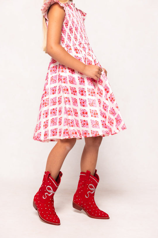 A girl stands against a plain white background, wearing a pink and white patterned dress with the playful Mini Channing Bootie in Red by Ivy City Co, creating an adorable mommy-and-me boots moment.