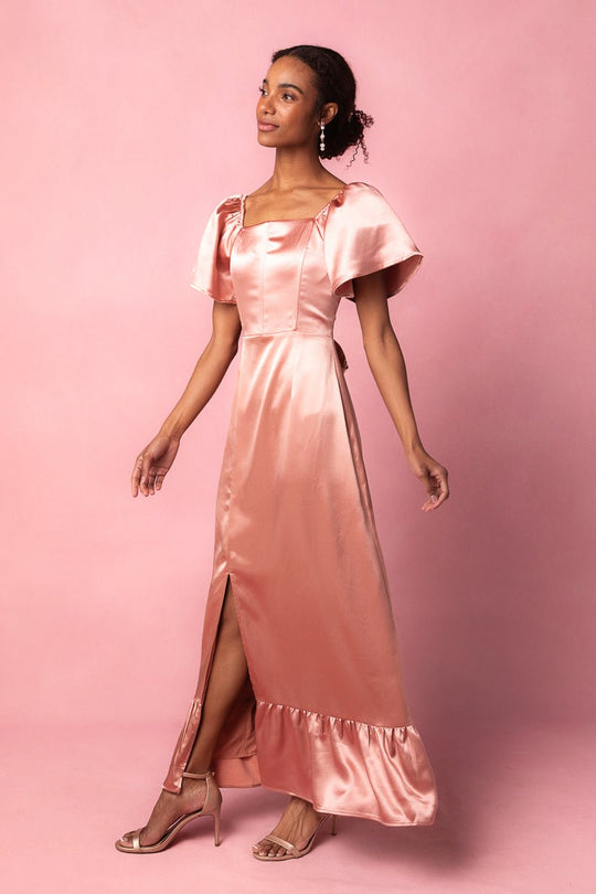 A woman stands against a matching pink background, looking to the side, wearing the Ivy City Co Maddie Dress in Apricot Crush Satin—a shiny, light pink short-sleeve dress featuring a square neckline and front slit. Styled with heels and dangling earrings, this look is perfect as a bridesmaid dress.