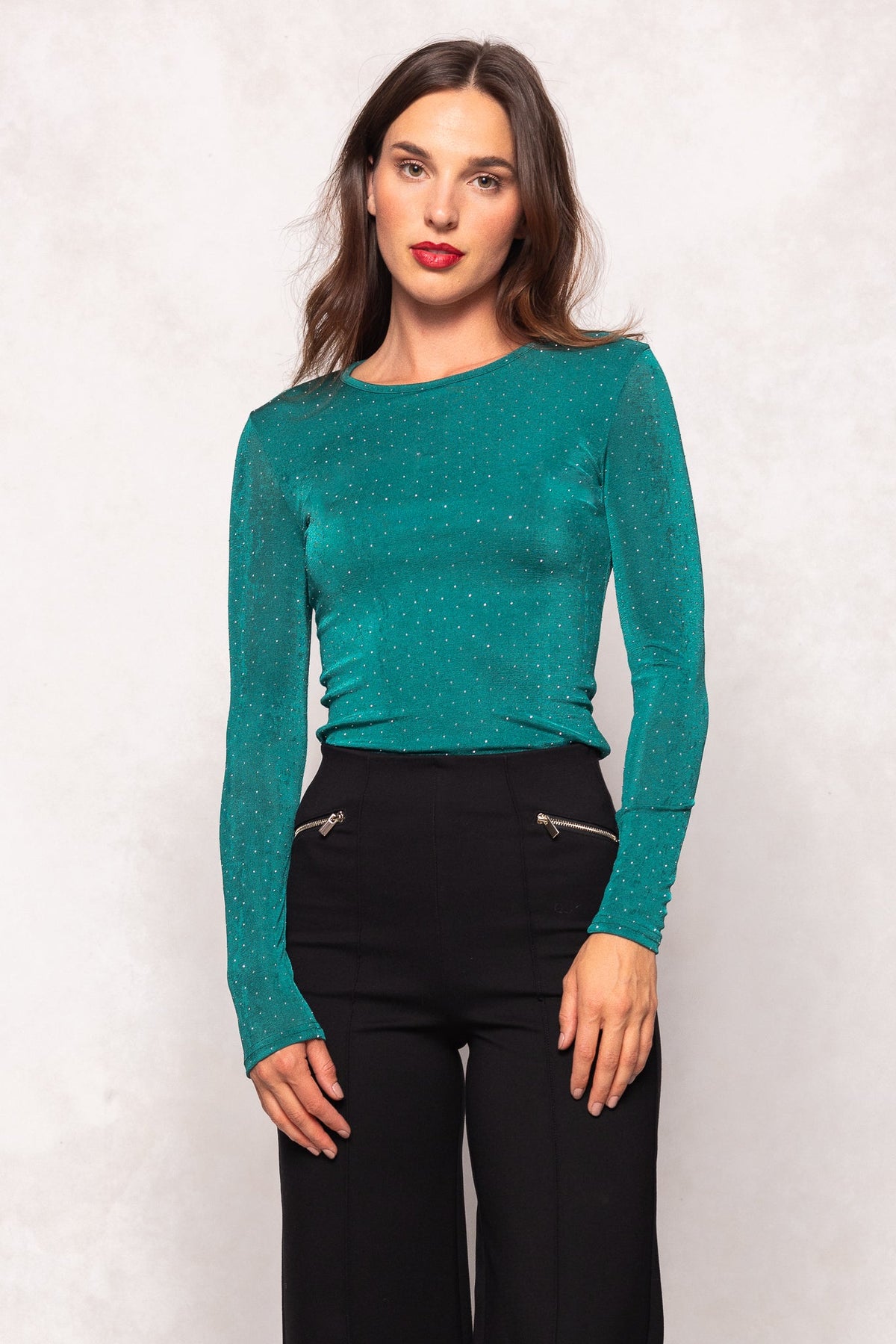 Woman with long brown hair wearing the Ivy City Co Bravo Top in Emerald—a maternity-friendly, teal long-sleeve top with small dots—paired with black high-waisted pants, standing against a light background.