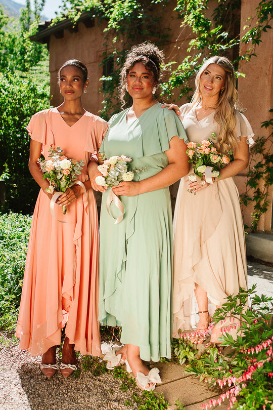 Three women, dressed in Ivy City Co’s Callie Dress in Champagne Chiffon, stand outdoors holding small bouquets and smiling. Greenery and sunlight surround them, with a vine-covered wall in the background.