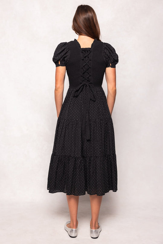 A woman with long brown hair, wearing silver flat shoes, stands with her back to the camera in the Ivy City Co Darby Dress in Black Diamonte. This stylish and maternity-friendly dress features short sleeves, small white dots, a lace-up back detail, and a tiered midi silhouette.