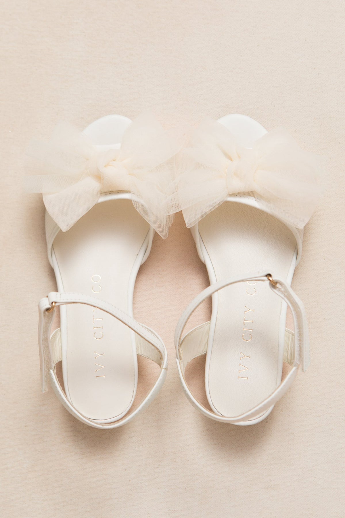 The Mini Ivy Bow Heel in White by Ivy City Co features comfortable white sandals with ankle straps and a large, soft tulle bow detail on the front. Displayed on a light beige surface, these shoes are perfect for any special occasion.