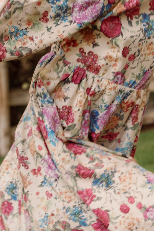 Close-up of the Mini Adelpha Dress in Autumn Amity Floral by Ivy City Co, a beige dress adorned with a vibrant floral pattern of pink, red, and blue flowers, shown outdoors. The moving fabric emphasizes the playful design and elegant smocked bodice, while balloon sleeves bring added volume and charm to this lively look.