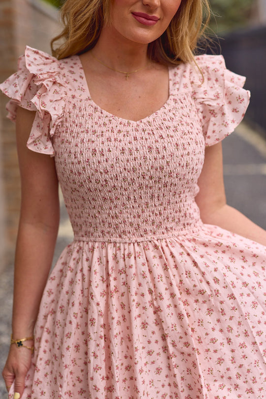 A woman wearing the Hattie Dress in Madeline Pink by Ivy City Co—a light pink, floral-patterned cotton midi dress with ruffled sleeves. Featuring a smocked bodice and a flowy waist, this dress is an ideal choice for a birthday girl. She stands outdoors, holding part of her dress.