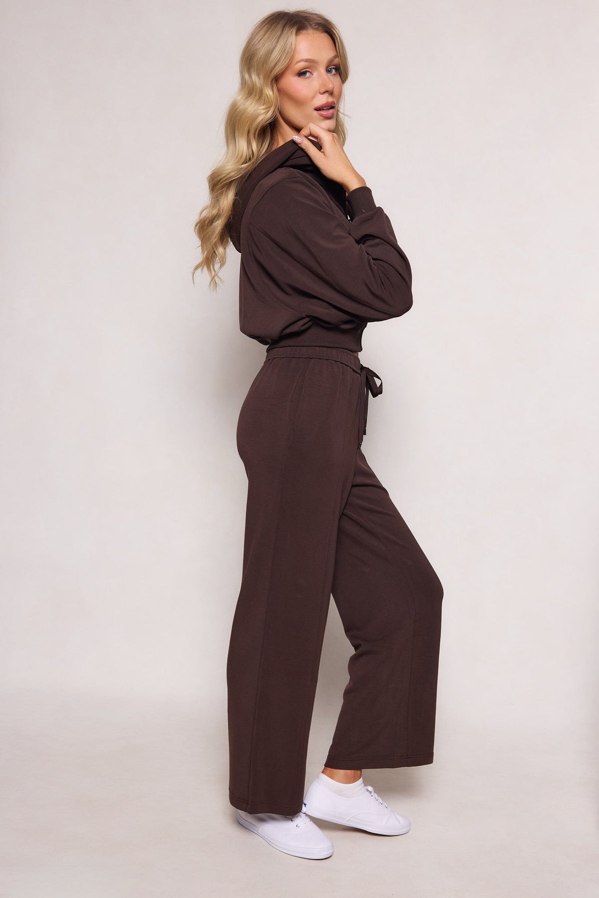 A woman wearing the Ivy City Co Amara Sweatpant in Espresso, styled with white sneakers, stands sideways against a light background. She has long wavy blonde hair and looks at the camera with her hand touching her chin.