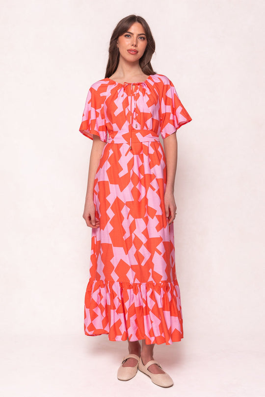 A woman wears the Ivy City Co Haven Dress in Geometric—a loose, ankle-length dress with bold orange and pink geometric patterns, flutter sleeves, and a ruffled hem—paired with beige slip-on shoes. She stands against a plain light background.
