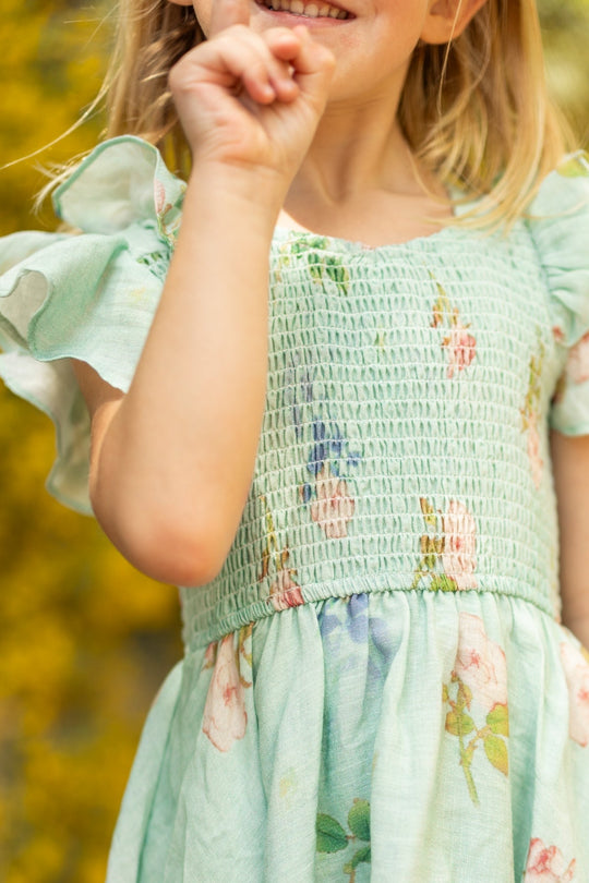 A young girl in the Ivy City Co Mini Hattie Dress in Roselyn Blue smiles and holds her hand to her face, standing outdoors with a blurred yellow background—a perfect choice for any girls party dress occasion.
