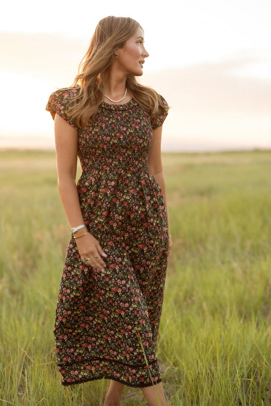 Dressed in Ivy City Co’s Annalise Dress in Obsidian Vintage Poppies, a woman stands amidst tall grass at sunset, her loose hair framing her face as she gazes to the side, accessorized with a necklace and bracelets. Maternity friendly and effortlessly chic, she radiates natural beauty against the glowing horizon.