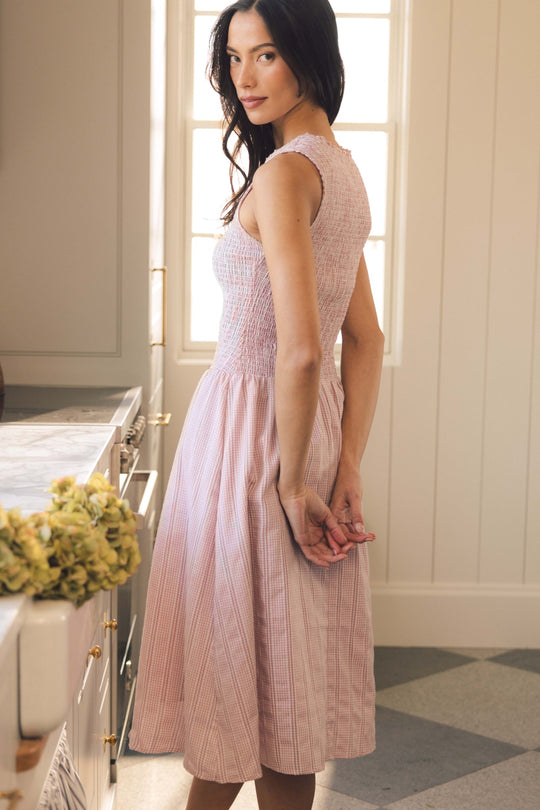 A woman wearing the Ivy City Co Karla Dress in Pink Striped Gingham stands in a sunlit kitchen, looking over her shoulder with her hands clasped behind her back. Yellow flowers sit on the counter in the foreground.